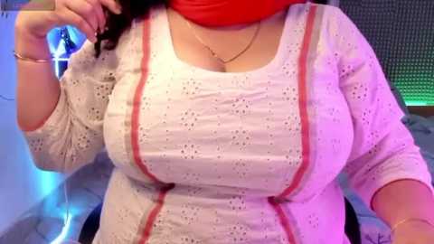 Media: Video of a curvy woman with light skin, large breasts, and a red hijab, wearing a white, lace-patterned top with red trim, sitting indoors with a blue and green LED light background.