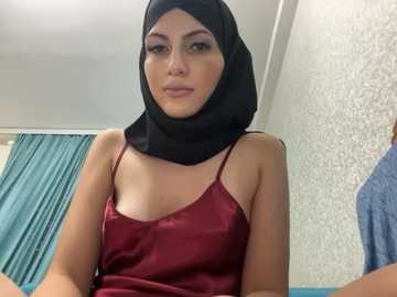 Media: Video of a woman with light brown skin, wearing a black hijab and a red satin camisole, sitting in a room with a teal curtain and textured white wall.