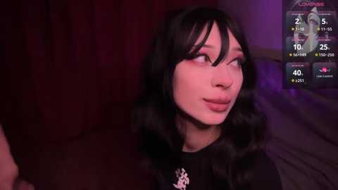 Media: Video of a young woman with long black hair and pale skin, wearing a black dress, in a dimly lit room with a purple glow.