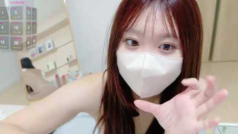 Media: Video of an East Asian woman with long, straight, auburn hair, wearing a white face mask, looking directly at the camera, in a clean, modern room with white walls and a calendar in the background.