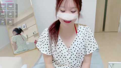 Media: Video of a young Asian woman with red hair in a ponytail, wearing a white polka-dotted dress and a face mask, sitting on a bed, with a large mirror reflecting her image.