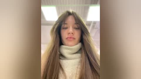 Media: Video of a young woman with long, straight, light brown hair, wearing a beige turtleneck sweater, in a brightly lit, modern indoor setting with fluorescent lights and white walls.