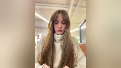 Media: Video of a young Caucasian woman with long, straight brown hair, wearing a beige turtleneck sweater, seated indoors in a modern, well-lit space with exposed ceiling beams and large windows.