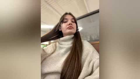 Media: Video of a young woman with long, straight brown hair, wearing a cream-colored turtleneck sweater, captured in a dimly lit, indoor setting with blurred background elements.