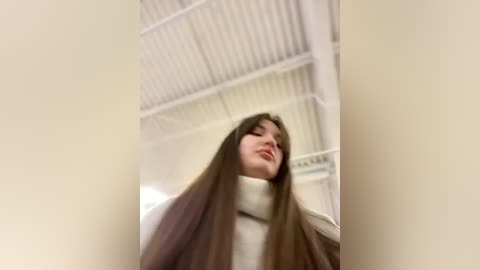 Media: Video of a young woman with long brown hair, wearing a beige turtleneck sweater, standing in a dimly lit room with white metal ceiling panels.