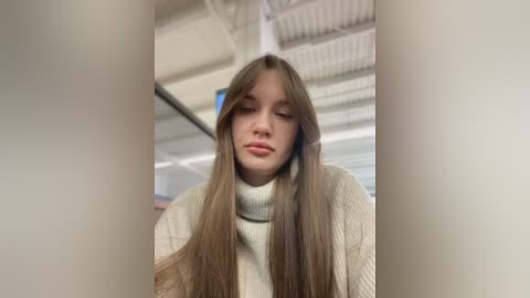 Media: Video of a young woman with long, straight brown hair, wearing a cream-colored turtleneck sweater, standing in a modern, well-lit indoor setting with white walls and visible ceiling beams.