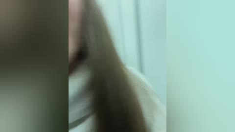 Media: A blurry video of a person with long brown hair, possibly a woman, in a dimly lit room with a white wall and a bed in the background. The image is out of focus, making it difficult to discern details.