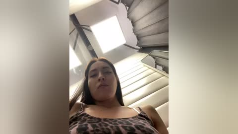 Media: Video of a young woman with light skin and long brown hair, wearing a leopard-print tank top, climbing a staircase. The background features a modern, gray, spiral staircase.