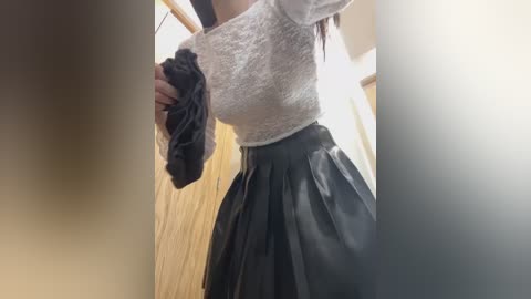 Media: Video of a person in a tight white knit sweater and pleated black leather skirt, holding black clothing against a wooden door and white wall background.