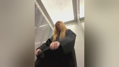 Media: Video of a blonde woman with long hair in a black coat, sitting on a narrow, white-tiled hallway floor, looking down, hands on knees.