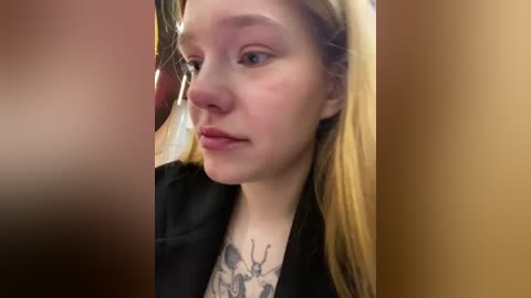 Media: Video of a young, fair-skinned woman with long blonde hair, wearing a black jacket and a floral shirt, with a deer tattoo on her chest.