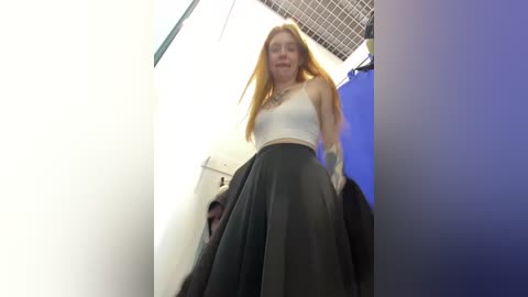 Media: A low-angle video of a young, fair-skinned woman with long, straight, blonde hair, wearing a white crop top and a black pleated skirt, standing in a brightly lit room with grid ceiling tiles and blue lighting.