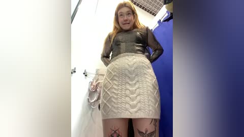 Media: Video of a young, fair-skinned woman with long, straight blonde hair, wearing a black mesh top and a beige, patterned, knee-length skirt, standing in a dimly lit, narrow hallway.