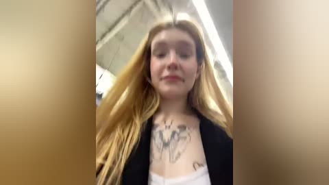 Media: Video of a young woman with long, straight blonde hair, wearing a black blazer over a white top, showing a tattoo of a mermaid on her chest.