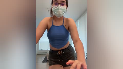 Media: Video of a young Asian woman with light skin, wearing a blue crop top, black shorts, and a surgical mask. She has glasses and is leaning forward, hands on her thighs, in a modern, bright room.