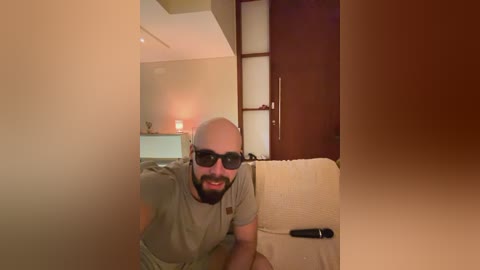 Media: Video of a bald, bearded man with sunglasses, wearing a beige T-shirt, sitting on a beige chair in a dimly lit room with wooden furniture and a lamp.