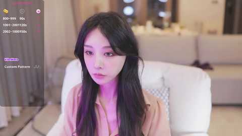 Media: Video of an East Asian woman with long black hair, wearing a beige top, seated in a modern living room with white couches and ambient lighting.