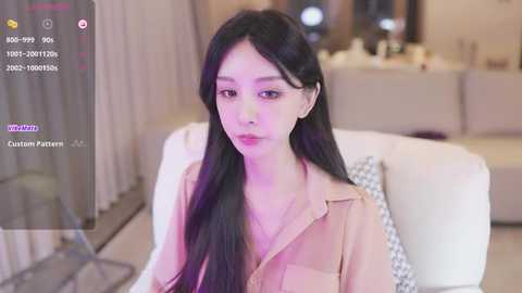 Media: Video of an East Asian woman with long black hair, wearing a beige button-up shirt, seated on a white couch in a modern living room with soft lighting and neutral tones.