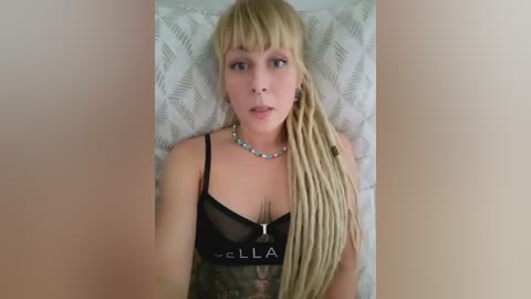 Media: A video of a fair-skinned, blonde-haired woman with long dreadlocks, wearing a black bra and a choker, lying on a bed with white, leaf-patterned sheets. She has a neutral expression.