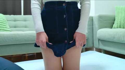 Media: Video of a woman in a white sweater and dark blue denim skirt, hands pulling the skirt up, revealing her bare thighs. Background includes light green sofas and a green throw pillow.