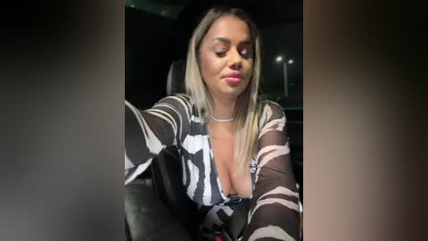 Media: Video of a light-skinned, blonde woman with a curvy physique, wearing a low-cut, patterned top, sitting indoors with a relaxed expression, eyes closed.