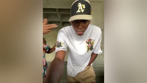 Media: Video of a young African-American man in a white t-shirt with colorful graphics, tan pants, and a black Oakland Athletics cap, reaching out in a gym locker room.