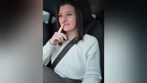 Media: Video of a woman with shoulder-length dark hair, wearing a white sweater, seated in a car with a seatbelt, making a shushing gesture, and smiling slightly.