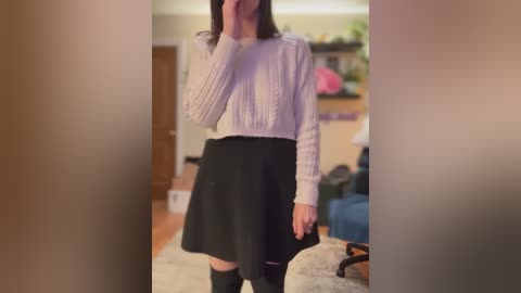 Media: Video of a slender woman with straight dark hair, wearing a white knit sweater and black skirt, standing indoors with blurred background.