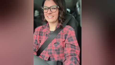 Media: Video of a smiling woman with short dark hair, wearing glasses and a red plaid shirt, seated in a car with a black seatbelt.