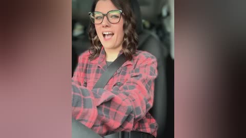 Media: Video of a woman with curly brown hair, wearing black-framed glasses, a red plaid shirt, and a black seatbelt, smiling and laughing in a car.