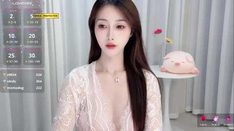 Media: Video of an Asian woman with long dark hair, fair skin, and delicate features wearing a lace, V-neck top. She sits in a modern, white room with a round table and a pink plush pig.