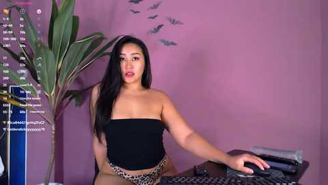 Media: Video of a young Asian woman with long black hair, wearing a black strapless top and leopard-print shorts, sitting at a desk in a purple room.