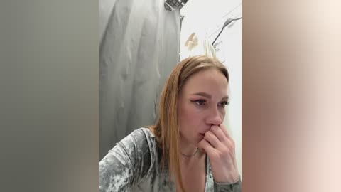 Media: A video of a blonde woman in a gray, patterned top, with her hand near her mouth, looking contemplative, in a dimly lit bathroom with a shower curtain and a light fixture in the background.