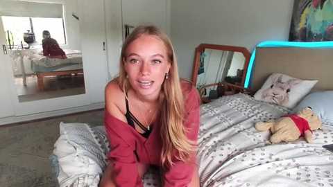 Media: Video of a blonde woman in a red cardigan, leaning forward on a bed with a teddy bear and a patterned duvet, in a modern bedroom with white walls and a mirror.