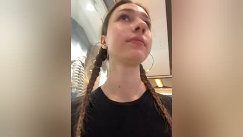 Media: A video of a young Caucasian woman with fair skin, brown hair in two braids, and a black top. She gazes upwards, possibly at a ceiling, in a dimly lit room.