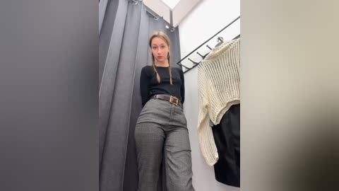 Media: Video of a young woman in a fitting room, wearing a black top, gray high-waisted pants, and a black belt. She has long blonde hair. Gray curtain, beige sweater hanging, and white walls.