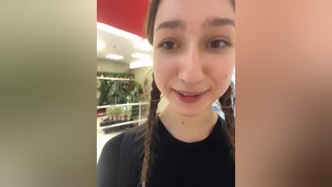 Media: Video of a smiling young woman with long brown hair in braids, wearing a black top, standing in a modern, brightly lit shopping mall with plants and shelves in the background.