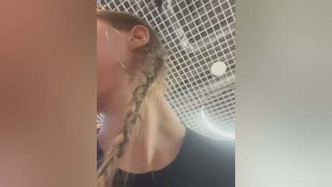 Media: Video of a woman with long, braided hair, wearing glasses and a black top, standing in a tiled shower with a large, circular showerhead visible.