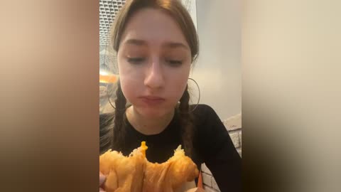 Media: Video of a young woman with straight, dark hair eating a golden-brown, deep-fried chicken wing, wearing a black top, in a kitchen with white cabinets and a window.