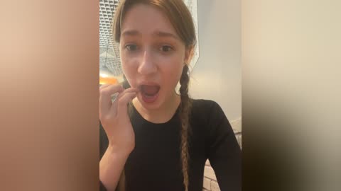 Media: Video of a young Caucasian woman with light skin and brown hair in braids, wearing a black top, licking her fingers in a playful manner, indoors with a beige tiled wall and a white curtain in the background.