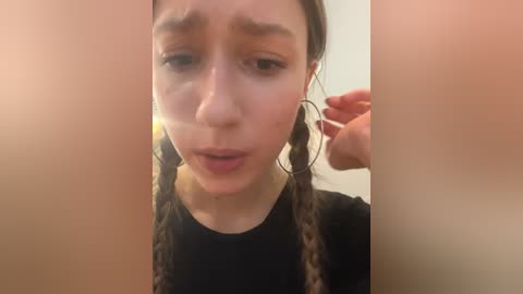 Media: Video of a young woman with light skin and dark brown hair in braids, wearing a black shirt. She has a neutral expression, and her hand is near her ear.