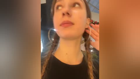 Media: A video of a young woman with fair skin, brown hair in braids, and large hoop earrings, standing indoors, wearing a black top, with a blurred background.