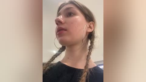 Media: A video of a young woman with fair skin, brown hair in braids, wearing large silver hoop earrings and a black top, taken from a low angle, highlighting her profile and expression.