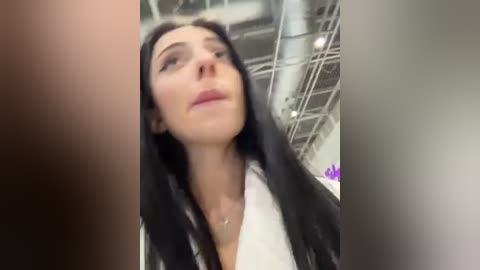 Media: Video of a young Caucasian woman with long black hair, fair skin, and light makeup, looking upward. She wears a white top. Background shows industrial ceiling and blurred text.