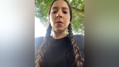 Media: Video of a young woman with long, dark hair in twin braids, wearing a black top, set against a green, leafy background.