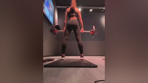 Media: Video of a muscular woman in a gym, wearing black leggings and a sports bra, squatting with a barbell on a gray mat, surrounded by dark gray walls and a large screen displaying a blue logo.