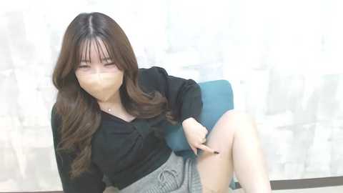 Media: Video of an Asian woman with long brown hair, wearing a black blouse and gray skirt, gagged with tape, sitting on a blue chair in a blurry, light-filled room.