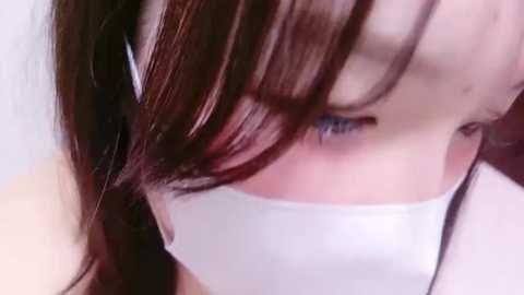 Media: Close-up video of a young Asian woman with long, dark brown hair, partially obscured by a white cloth gag. Her fair skin and blue eyes are visible, and she appears distressed, suggesting a potentially distressing or threatening situation.