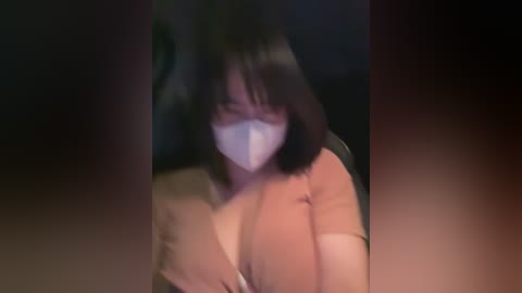 Media: A blurred video of a woman with short black hair and a white mask, wearing a light-colored top, seated in a dark, dimly-lit room.