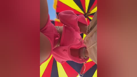 Media: A close-up video of a person wearing a bright pink, frilly dress with lace details, standing under a vibrant, multi-colored umbrella with red, yellow, and black stripes.